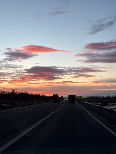The image, oriented vertically but depicting a horizontal landscape, captures a serene yet dramatic sunset or sunrise scene from the perspective of someone traveling on a multi-lane highway.

The left half of the image is dominated by a magnificent sky, transitioning from a soft, pale blue at the top to a vibrant tapestry of orange, pink, and yellow closer to the horizon. Streaky, elongated clouds are painted with intense hues of red and orange, particularly in the lower and middle portions of the sky, contrasting with pockets of lighter white and gray clouds. The overall effect suggests the magical hour of twilight.

The right half of the image features a dark asphalt highway, seemingly stretching into the distance. White lane markings clearly delineate the travel lanes and a shoulder on the right side. The road surface appears dark, possibly due to low light conditions or a slight dampness, reflecting minimal light.

Several vehicles are visible on the highway. In the mid-ground, a dark silhouette of a large vehicle, possibly a truck or a semi-trailer, is prominent against the brighter horizon. Further down the road, a second, lighter-colored vehicle, which appears to be a semi-truck, is clearly distinguishable with its yellow or amber headlights glowing softly, illuminating the path ahead. In the far distance, faint pinpricks of light indicate other vehicles.

Along the horizon, separating the colorful sky from the highway, is a dark, silhouetted landscape. This appears to be a treeline or dense vegetation bordering the road. On the left side of the highway, a series of utility poles or similar structures are visible in silhouette against the vibrant sky.

The time of day is clearly either sunrise or sunset, given the striking colors in the sky and the low ambient light, which necessitates the use of vehicle headlights. The atmosphere is calm and contemplative, emphasizing the beauty of nature even amidst infrastructure like a highway. No discernible text is visible in the image.