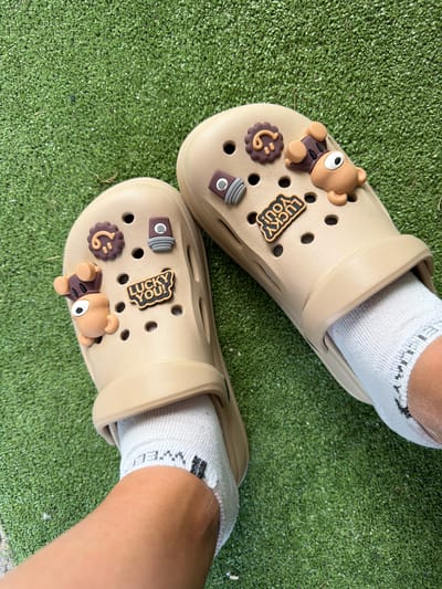 A pair of tan-colored Crocs, adorned with cartoonish charms of a bear, a flower, a spray can, and the words "LUCKY YOU!", are worn by an individual whose feet, clad in white socks, rest on a patch of artificial turf. The scene is outdoors, with the artificial turf providing a green, textured ground cover. The lighting suggests it is daytime. No specific location in the city of Rusokastro, Bulgaria, can be identified from the image.