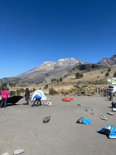 The image displays an outdoor scene in Tlamacas, Mexico, under a clear, bright blue sky. A large mountain range, partially snow-capped or featuring light-colored rock near its peaks, dominates the background, with lower slopes exhibiting sparse, dry vegetation and scattered pine trees. The midground consists of an undulating landscape covered in yellowish-brown grass and occasional small trees. The foreground is a flat, dark grey, gravelly or dirt surface.

Three individuals are present in the left-center. One person, wearing a blue jacket and dark pants, is bent over, facing away from the camera, appearing to be working on a light blue dome tent that is partially erected or being disassembled. To the left of this person, another individual in a dark (black) jacket and dark pants stands with their back to the camera, observing. Further left, a third person in a bright pink jacket and dark pants stands facing towards the center of the scene.

Various objects are scattered on the ground: a large dark tarp lies partially unfolded to the left of the individuals. An orange sleeping bag or rolled blanket is spread out on the ground to the right of the tent. A dark green duffel bag or backpack rests in the central foreground. Two smaller blue tarps or pieces of fabric are on the ground in the foreground-right. Scattered rocks, some forming a loose circle, are visible around the light blue tent. In the far right, a white SUV is partially visible, parked near a series of wooden posts and a green information sign.