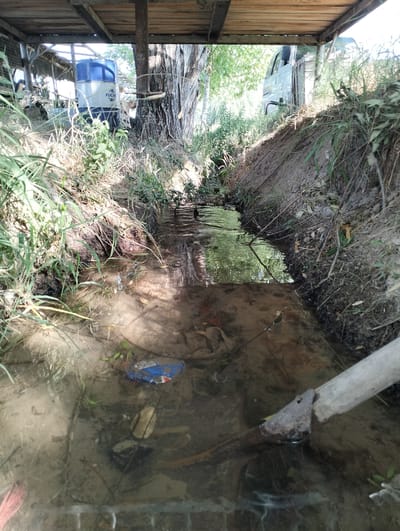 The image depicts a shallow, murky stream flowing through a rural or semi-rural environment. The stream banks are eroded, with exposed soil and vegetation. Debris, including a crumpled blue and silver object, is visible in the water. Overhead, a wooden structure with beams is present, partially obscuring the sky and surrounding trees. In the background, glimpses of a white vehicle and a blue and white cylindrical container with the logo "FUCHS" are visible. The overall scene suggests an outdoor area, possibly near a farm or workshop, in Aktam (Tashminvody), Uzbekistan.