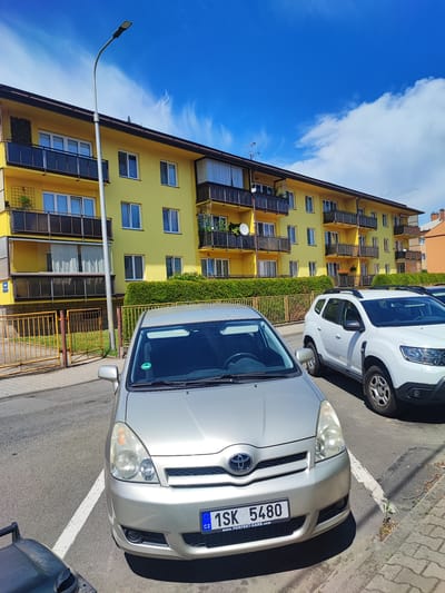 A silver Toyota Verso car is parked in a parking space in front of a bright yellow apartment building. The car has a Czech license plate that reads "1SK 5480" and "WWW.PERFEKT-CARS.COM". To the right of the Toyota, a white Dacia Duster is parked. The apartment building has several balconies with dark railings and appears to be multi-story. A street lamp is visible on the left side of the frame. The scene is set on a sunny day with a bright blue sky and some wispy white clouds. A green hedge runs along the front of the apartment building, and a yellow metal fence is visible behind the parked cars. The image appears to be taken in Nový Jičín, Czechia.