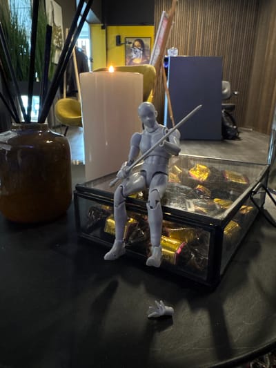 An interior scene in Ålesund, Norway, presents a dark tabletop displaying a collection of objects. Centrally positioned, a gray articulated human-like figurine sits on a transparent glass box with dark metal framing. The box contains numerous individually wrapped candies in gold and red foil, with "Werther's Original" visible on some wrappers. The figurine holds a slender, light-gray rod diagonally across its torso. To the lower right of the figurine, a detached gray hand, matching the figurine's material, rests on the table surface. Behind the figurine, a tall, white cylindrical candle with an active flame casts light. To the left, a dark brown ceramic vase holds several long, dark reed diffusers or decorative sticks. The dimly lit background, out of focus, reveals sections of a bright yellow wall with a framed artwork depicting a masked face, a vertically textured wall, a light-colored easel, a dark blue rectangular panel, a black bag, and a yellow upholstered chair, suggesting a commercial or lounge setting.
