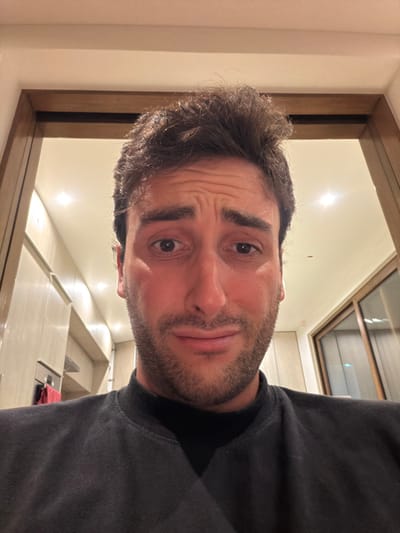 A light-skinned man with short, dark curly hair and a trimmed beard gazes at the camera. He has brown eyes and slightly furrowed brows. He wears a dark-colored sweater. The background reveals a well-lit interior, likely a kitchen, with a reflective countertop and a skylight visible above. The image is slightly rotated. The man is centered in the frame.