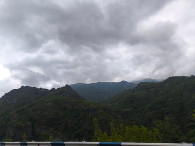 The image shows a view of lush green mountains under a cloudy sky. In the foreground, there is a white barrier with blue and red stripes. The mountains are densely covered with trees and vegetation, with some rocky outcrops visible on the left. The sky is overcast with dark gray clouds, suggesting a possibility of rain or an impending storm. The scene appears to be a natural landscape, possibly a roadside view, in the vicinity of Aygehat, Armenia, a region known for its mountainous terrain and natural beauty. There are no people or animals visible in the image. The overall impression is one of a dramatic and somewhat brooding natural landscape.