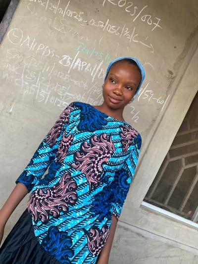 A young woman wearing a light blue head covering and a blue, pink, and black patterned top stands against a wall covered in handwritten text. The text appears to be a mix of numbers and abbreviations. The woman looks at the camera with a slight smile. A window or opening is visible in the upper right corner of the image. The photograph is taken at an angle. The woman is wearing a black skirt or pants.