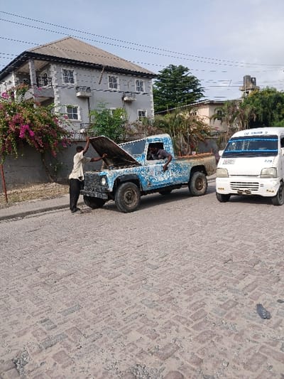 The scene shows two men inspecting a blue, Land Rover Defender parked on a street in Way, Nigeria, which is located at 6.429° N, 3.468° E. The Defender has its hood open and is facing to the right. The street is paved with red bricks, which are cracked and worn down. A white commercial van with its windows rolled up, is parked to the right of the Defender. The van is facing to the left. The van has "God Knows" written on the side. There is a gray, two-story house to the left of the image, with a yard containing shrubs. The house has a black roof and a white concrete wall. The house has windows on the upper story and the ground level. There are power lines and poles running above the street. The scene is taken from a distance and at a slight angle. The weather appears to be clear. The image shows a common scene in Nigeria, where people are often seen repairing vehicles on the side of the road.