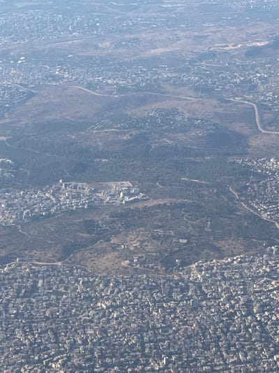 This aerial photograph captures a dense urban landscape, characteristic of Rosh HaAyin, Israel. The image displays a mix of residential and possibly commercial areas, with a vast expanse of tightly packed buildings dominating the foreground. The architecture appears to be predominantly low-rise to mid-rise, with many structures featuring light-colored facades and flat roofs, common in the region.

Further into the mid-ground, the urban density gradually thins out, giving way to patches of undeveloped land, sparsely vegetated hillsides, and a significant forested area. Winding roads and pathways are visible throughout the landscape, connecting different parts of the city and the surrounding natural terrain. In the distance, the urban sprawl continues, merging with the hazy horizon, suggesting a larger metropolitan area or continued development.

The overall impression is one of a sprawling city situated within a hilly, somewhat arid environment, typical of the Israeli landscape. The lighting suggests it's daytime, with shadows indicating the sun's position, though the haziness common in aerial shots slightly softens the details. There is no visible human activity or specific objects of interest that stand out, presenting a general view of urban development and its integration with the natural surroundings.