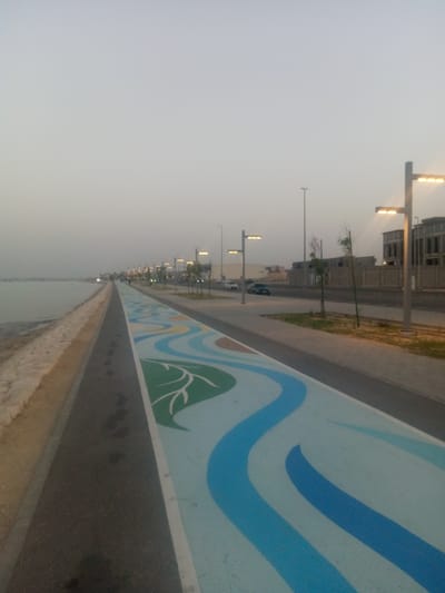 The photograph depicts a waterfront pathway in Al Khobar, Saudi Arabia. The pathway is divided into two sections: a darker gray asphalt area along the water's edge and a lighter-colored, decorated lane adjacent to it. The decorated lane features a design of blue waves and green leaf-like shapes on a pale blue background. Footprints are visible on the darker asphalt path.
Parallel to the pathway is a road with streetlights evenly spaced. Beyond the road, there are sidewalks, sparse vegetation, and buildings under construction or recently completed. Several vehicles are parked along the road. The sky is overcast, suggesting dusk or dawn.
