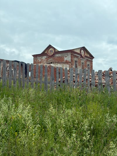The image captures a dilapidated brick building with a weathered wooden fence in the foreground, partially obscured by lush green vegetation and wildflowers. The building's facade shows signs of decay, with exposed bricks and missing windows. Two circular decorative elements are visible on the upper part of the building, one on the gable and another higher up, both appearing to contain painted images. The fence is constructed from rough-hewn wooden planks, some of which are broken or missing, contributing to the overall impression of neglect. Dense green foliage and numerous small white flowers dominate the right side of the frame, creating a stark contrast with the crumbling architecture. The sky above is overcast with a pale, diffused light, suggesting a cloudy day, possibly morning or afternoon. The scene conveys a sense of abandonment and the passage of time, with nature reclaiming the man-made structure. The location is specified as "Юг, Russia." No visible text is present in the image.