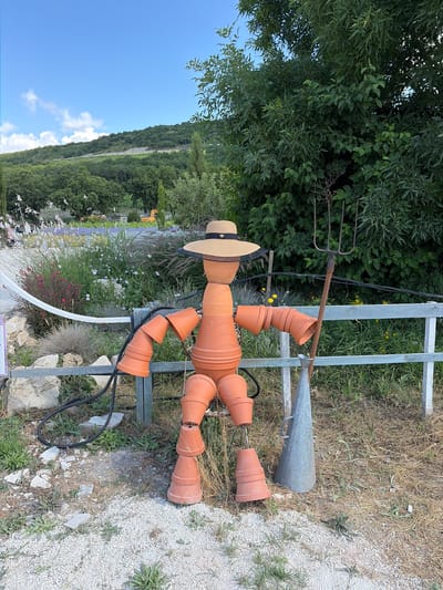 The image shows a scarecrow constructed from terra cotta pots, standing in a garden setting. The scarecrow is wearing a straw hat adorned with a bee emblem. It is holding a garden fork and a metal funnel. The background features lush greenery, flowering plants, and a hilly landscape under a clear blue sky with scattered clouds. The ground is covered with gravel and dry grass. The scene is set in Semogorskiy, Russia.