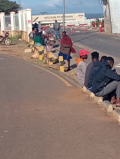 This image depicts an outdoor scene at the Namanga One Stop Border Post, located in Namanga, Kenya, with signage welcoming visitors to Tanzania. The setting appears to be a sunny day, likely morning or late afternoon, under a clear blue sky with some scattered clouds.
In the foreground and to the right, a paved road runs alongside a curb made of yellow and black striped concrete blocks. A group of approximately four to five young men are seated on this curb, appearing to be waiting. One wears a red baseball cap and an orange hoodie, while another is in a dark jacket. Their posture suggests a relaxed, patient demeanor.
Further into the scene, closer to the main border infrastructure, several other individuals are visible. A prominent figure is a man in the mid-ground wearing a traditional red plaid garment (resembling a Maasai shuka) over blue shorts, holding a stick. Nearby, another person is seen in green attire with a head covering. More people are gathered closer to the border checkpoint, some carrying bags.
To the left, a light-colored building with metal fencing and a red motorcycle parked beside it is visible. The border itself is marked by red and white striped barrier arms. A large sign in the background clearly reads: "WELCOME TO TANZANIA" and "NAMANGA ONE STOP BORDER POST". The overall scene suggests a typical day at a busy land border crossing, with people waiting to cross.