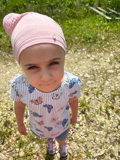 A young girl wearing a pink headscarf and a blue and white striped t-shirt with butterflies is standing outdoors in what appears to be a rural or park-like setting. The ground is covered in small stones and sparse vegetation, and the background consists of green grass and some wooden structures. The girl is looking directly at the camera with a neutral expression. She is wearing blue denim shorts and pink and gray sneakers.