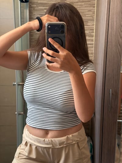 A young woman with long, dark brown hair is taking a mirror selfie in what appears to be a bathroom. She is wearing a black and white striped crop top and light beige pants. Her left hand is raised, adjusting her hair, and her right hand holds a smartphone, obscuring her face. She is wearing a black hair tie on her left wrist. The background shows bathroom tiles, a shower door, and a wooden cabinet.