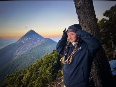A person stands in front of a mountain range. The person is wearing a dark jacket, a brown hat, and has blonde braids. Their hands are raised near their head, seemingly adjusting their hood. The person is leaning against a tree. In the background, a large mountain rises above the landscape, with other mountains and green foliage visible. The sky has hues of blue, orange, and white, suggesting either sunrise or sunset.
The location is not Placencia, Belize.
