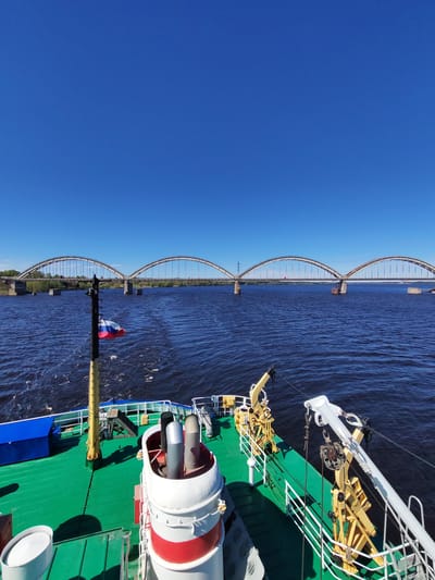 The image depicts a view from the deck of a vessel traveling on a river near the city of Zarya, Russia. A multi-arched bridge spans the river. The Russian flag is visible on a pole attached to the ship. The deck of the vessel is green, with some blue structures visible. The environment includes a blue sky, a river with visible currents, and the bridge. There are no people visible. Several pieces of ship equipment are visible on the deck.