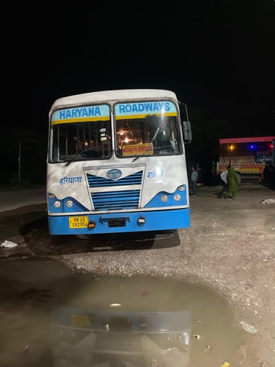 A Haryana Roadways bus is parked at night in Katauhar Kalan, India. The bus, white with blue accents, has "HARYANA ROADWAYS" printed in blue across its front window. A license plate reading "HR 45 GV2954" is visible on the front bumper. The bus is parked on a gravelly surface with puddles of water reflecting the bus and the night sky. In the background, other vehicles are present, including a truck with colorful lights and a few people standing near it. The overall scene suggests a bus stop or a roadside resting area at night.