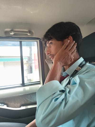 A young man with dark hair and a light blue collared shirt is seen from the chest up, looking out the window of a vehicle. He has his right hand raised to his ear. A dark lanyard with the words "Government College" and "Ramshala" is visible around his neck. The interior of the vehicle appears to be a light gray color. The window shows a blurred exterior with buildings and what appears to be a street scene. The image is likely taken in Sidhpur, India, given the context provided about the "Government College Ramshala."