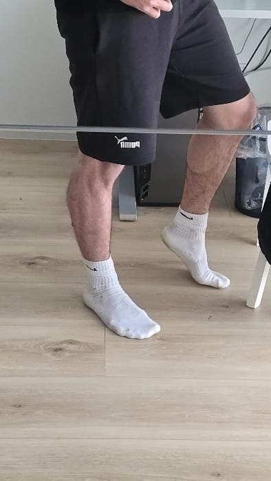 The image shows the lower half of a person wearing black Puma athletic shorts and white socks. They are standing in a room with light wood laminate flooring and a partial view of a white table and chair to the right.  The person's left leg is bent at the knee with their foot flat on the floor. The right leg is straight with the foot resting on the edge of the table. The image was taken in a room in Großkarolinenfeld, Germany, located at latitude 47.893, longitude 12.088.