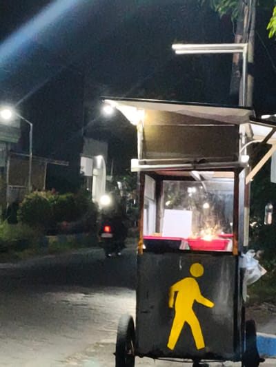 The image depicts a nighttime scene on a street in Sidorejo, Indonesia. A food cart, illuminated by a bright light at the top, is prominently featured in the foreground. The cart has a dark-colored base with a yellow pedestrian silhouette painted on its side. Inside the cart, a red tray is visible. To the left of the cart, a motorcycle with its headlights on is partially visible, along with some green foliage. In the background, streetlights cast a glow on the dimly lit road and surrounding structures. The overall atmosphere is urban and nocturnal.