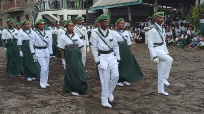 The image shows a group of people in uniform marching in a parade. They are all wearing white pants, white shirts, green skirts, and green hats. The people are marching in a line on a dirt path. The image was taken in Keshero, Democratic Republic of the Congo at latitude -1.624 and longitude 29.157. There are other people watching the parade in the background. There is a building in the background as well. The people in the background are wearing various clothing, including white shirts, green skirts, green pants, and green hats. They are sitting and standing and watching the parade. There is a small hill in the background to the left of the building. The ground is dirt and there are small plants growing on the ground. There are trees in the background. There is a sign above the building in the background.
