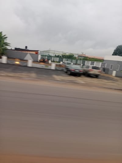 A low-angle, slightly blurred shot captures a street scene in Umuosonwu, Nigeria. The foreground is dominated by a wide, asphalt road, appearing somewhat out of focus due to motion blur. Beyond the road is a paved area with dirt and gravel, leading up to a low, grey building with white pillars. This building has dark windows and appears to be a commercial or residential property. A tall, dark water tank sits atop the building's roof. To the left, a lush green palm tree adds a touch of vegetation. In front of the building, a black car is parked, with a lighter-colored SUV parked behind it. A black fence separates the property from the street. In the background, a large, green net structure, possibly a sports court, is visible. The sky is overcast and grey, suggesting a cloudy or possibly rainy day.