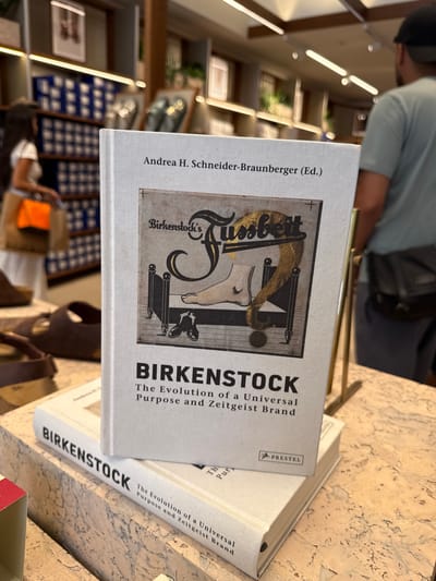 The image captures a display of Birkenstock books inside a retail store. In the foreground, two books are stacked. The top book, standing vertically, displays the title "BIRKENSTOCK: The Evolution of a Universal Purpose and Zeitgeist Brand" with the name "Andrea H. Schneider-Braunberger (Ed.)" above. A graphic featuring a foot on a bed is visible on this book's cover. Below it, another book lies horizontally with "BIRKENSTOCK" embossed on its spine and the subtitle "The Evolution of a Universal Purpose and Zeitgeist Brand" on its cover. The books rest on a textured surface.

In the background, out of focus, shelves are stocked with what appear to be shoeboxes, possibly Birkenstock products. A person wearing a light-colored t-shirt and dark pants is visible on the right, facing away from the camera. On the left, a woman with long dark hair wearing a white top and a beige skirt is browsing, holding an orange bag. The store's interior features bright lighting and wooden accents.

The scene is consistent with a retail environment in a Birkenstock store. The presence of Birkenstock merchandise and promotional material, combined with shoppers, indicates a commercial setting. The city of La Roca del Vallès, Spain, is known for its large outlet shopping center, "La Roca Village," which houses numerous brand stores including Birkenstock. Therefore, this image likely depicts a Birkenstock store within La Roca Village, La Roca del Vallès, Spain.