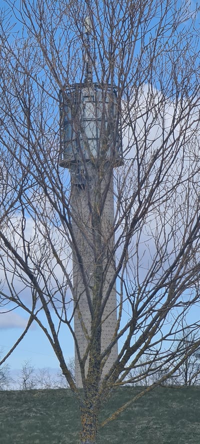 The image depicts a tall, rectangular structure behind bare tree branches. The structure has a light gray or white facade with a grid-like pattern visible on its surface, possibly brickwork. A rectangular object with glass panels and a metal frame is attached to the facade. The sky is visible through the branches, with patches of blue and white clouds. The scene is likely in Salaspils, Latvia.