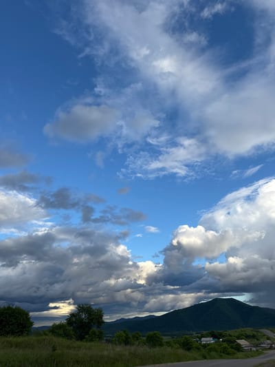 This is a vertical landscape photo with a dramatic sky filled with a mixture of bright white and dark gray clouds against a vibrant blue sky. The clouds are dynamic, with some appearing wispy and scattered, while others are dense and turbulent, suggesting changing weather. Sunlight is breaking through in some areas, creating highlights on the cloud formations.
In the lower right portion of the image, a lush green hillside is visible, dotted with trees. A rough, unpaved road runs along the top of the hill. Further down the slope, there are a few simple, rustic buildings with visible roofs, suggesting a rural or semi-rural settlement. The overall scene evokes a sense of raw natural beauty and a remote, tranquil setting.
The time of day appears to be late afternoon or early evening, given the angle of the light and the dramatic cloud formations. The weather is dynamic, with elements of both sunshine and potential storm clouds present. The location cues, such as the style of buildings and the type of vegetation, along with the provided context of Теплоозёрск, Russia, point to a location in a mountainous or hilly region of Russia.
The image does not contain any visible text. The overall mood is one of awe inspired by the power and beauty of nature, with a hint of the quiet life of a rural community.
