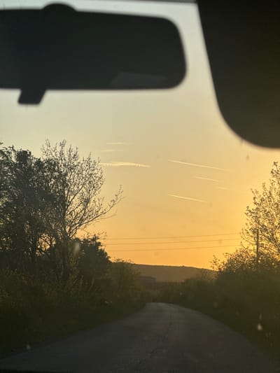 The photograph, taken from inside a vehicle, presents a view of a landscape under an orange-tinted sky. In the sky, several contrails from aircraft are visible. Power lines stretch across the upper portion of the frame. The foreground features the silhouette of the vehicle's interior, while the middle ground is occupied by trees and vegetation. To the right, a road is seen. The horizon reveals a low hill or mountain range.
There is no indication this image is in Gromshin, Bulgaria.