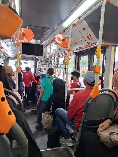 Inside a public transport bus, several passengers are visible. A man in a red shirt and jeans sits towards the front. Behind him, a person wears a green shirt. Overhead, the bus interior features numerous orange hand grips attached to the ceiling, along with white horizontal bars. Windows line the upper portion of the bus walls, letting in light. The scene depicts a typical commute with people seated and standing.