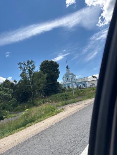 This image depicts a serene scene in Глебовское, Russia. The primary subject is a beautiful blue and white church with a prominent dome and spire, set against a bright blue sky dotted with wispy white clouds. The church appears to be an Orthodox Christian place of worship.
The setting is rural, with lush green trees surrounding the church and a dirt road running alongside it. The road is bordered by a strip of grass and vegetation. In the foreground, there's a portion of what appears to be a vehicle's interior, likely the source of the angled perspective, suggesting the photograph was taken from a car.
The time of day is clearly daytime, with strong sunlight illuminating the scene and casting shadows. The weather appears pleasant and clear, ideal for outdoor activities or sightseeing. There are no people visible in the frame, emphasizing the peaceful and quiet atmosphere.
Notable details include the architectural style of the church, which suggests a traditional Russian design. The contrast between the vibrant blue of the sky and the light blue and white of the church is striking. The overall impression is one of tranquility and the beauty of the Russian countryside. No visible text is present in the image.