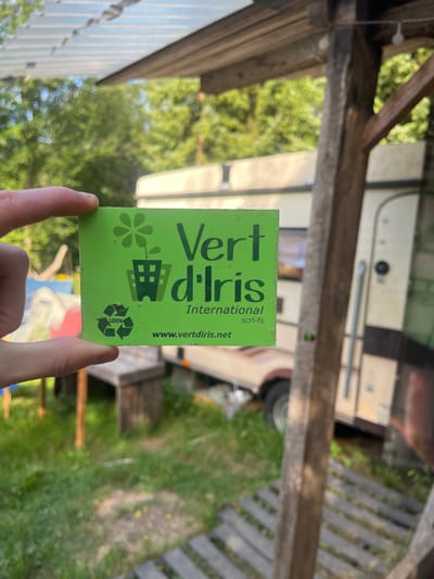 The image depicts an outdoor scene in Sint-Pieters-Leeuw, Belgium, under clear daylight conditions. In the foreground, a light-skinned hand holds a rectangular, bright green card or sticker. The card displays the dark green text "Vert d'Iris" prominently, with "International" and "scrl-fs" below it, and "www.vertdiris.net" at the bottom. A logo on the left side shows a stylized flower growing from a building, and a "100%" recycling symbol is present.
The background is an out-of-focus outdoor environment. A beige or off-white caravan/trailer with dark trim is parked on green grass directly behind the card. Parts of a rustic wooden structure, including a vertical support post on the right and horizontal beams overhead, are visible, suggesting a pergola or shelter. Further in the background, dense green foliage from trees forms a blurred backdrop. A wooden bench is partially visible on the ground to the left of the caravan.