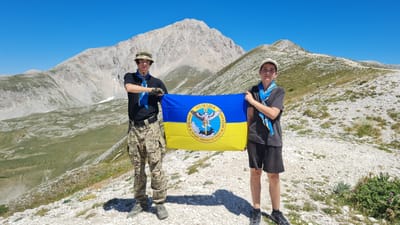 Two young males, appearing to be teenagers, stand on a rocky, mountainous terrain under a clear blue sky. The male on the left wears a camouflage hat, a black t-shirt, camouflage pants, and a blue scarf. He has his arms crossed and is looking towards the camera. The male on the right wears a white cap, a dark grey t-shirt, dark shorts, and a blue scarf. He is also looking towards the camera. Between them, they hold a Ukrainian flag, which is horizontally divided into blue and yellow sections, with a circular emblem in the center. The emblem features an eagle with outstretched wings, holding a sword and a shield, and text in Ukrainian and Latin.

The background consists of rugged, light-colored mountains with sparse green vegetation in the lower valleys. The terrain is characterized by gravel and rocks. The overall environment suggests an elevated, possibly remote, natural setting. The location is identified as the Gran Sasso mountain range, near L'Aquila, Italy.