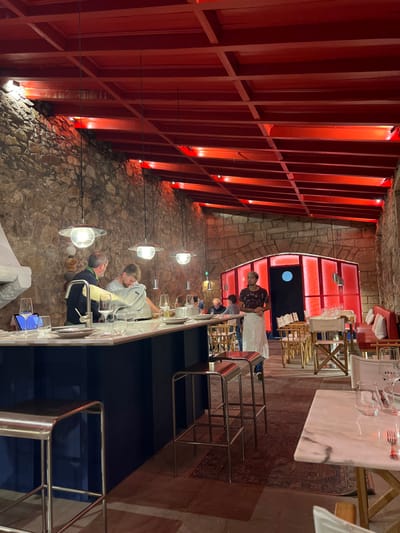 The image displays the interior of a restaurant or bar located in Caminha, Portugal. The space features rough, exposed stone walls and a modern, red-painted slatted wooden ceiling, which is illuminated by integrated red uplighting. Multiple modern pendant lights, characterized by a saucer-shaped top and a clear spherical globe, are suspended over a dark blue counter with a white marble-patterned surface.
On the counter, a tablet in a stand, several wine glasses, a silver-toned faucet, plates, and a commercial deli slicer are visible. Behind the counter, two men are present: one in a dark sweater observes the second man, who is operating the deli slicer. To the right of the counter, a male server, wearing a dark patterned shirt and a white apron, stands attentive.
The dining area to the right features wooden tables with matching chairs, and a red banquette with white cushions lines the far wall. A distinct, red-illuminated, translucent hexagonal structure with a circular dark opening is positioned in the background, possibly functioning as a service area or enclosed space. The floor is covered with what appears to be stone or concrete, partially accented by a patterned rug. Several patrons are seated at tables throughout the establishment.