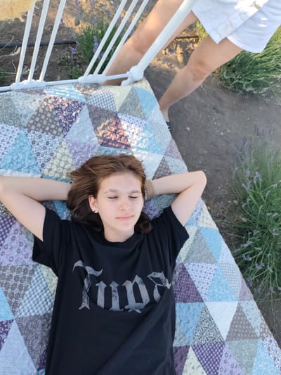 A young person with brown hair is lying on their back on a patchwork quilt. They are wearing a black t-shirt with a gray gothic-style text graphic. Their hands are clasped behind their head, and their eyes are closed, suggesting a relaxed or contemplative mood. The quilt is spread out on the ground, which appears to be dirt or gravel. In the background, there is a white metal structure, possibly a bed frame or a bench, and tall green plants with small purple flowers, characteristic of lavender. Another person's legs, wearing white shorts, are visible at the top of the frame, suggesting they are standing near the person on the quilt. The lighting suggests it is daytime, possibly late afternoon or early evening, given the warm tones. The setting appears to be outdoors, in a field or garden with lavender plants. The visible text on the t-shirt is gothic-style lettering.