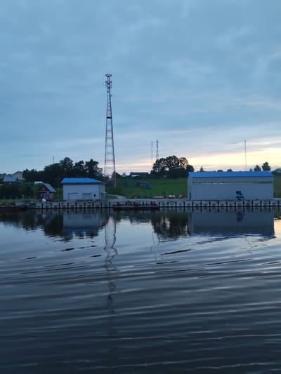 The image depicts a tranquil scene at the edge of a body of water, likely a river or canal, in Ivanov Bor, Russia. The time appears to be dusk, as indicated by the soft, diffused light and the muted colors of the sky. In the foreground, the calm water reflects the structures on the shore and the sky above, creating a mirror-like effect. The water is rippled, suggesting a gentle breeze or the wake of a passing boat.
Along the shoreline, a concrete quay or embankment is visible, lined with what appear to be pilings or mooring posts. Several buildings are situated along this embankment. The most prominent is a long, modern-looking industrial or warehouse-style building with a blue, corrugated metal roof and grey, ribbed metal walls. To the left of this building is a smaller, older structure with a white facade and a dark-colored roof, possibly a house or an office. Further to the left, partially obscured by trees and a gentle slope of green grass, are several other residential-style houses with pitched roofs, suggesting a small settlement or village.
Dominating the skyline and serving as a focal point is a tall, lattice-style transmission tower, likely for telecommunications or broadcasting. Its red and white sections are clearly visible against the pale blue and grey sky. Two other, shorter towers, also appearing to be transmission masts, are visible further in the background. The sky itself is a blend of soft blues and greys, with hints of warmer tones near the horizon, suggesting the last vestiges of sunset. The overall impression is one of a quiet, functional industrial area situated within a natural, possibly rural, setting. There are no people or animals visible in the frame.
