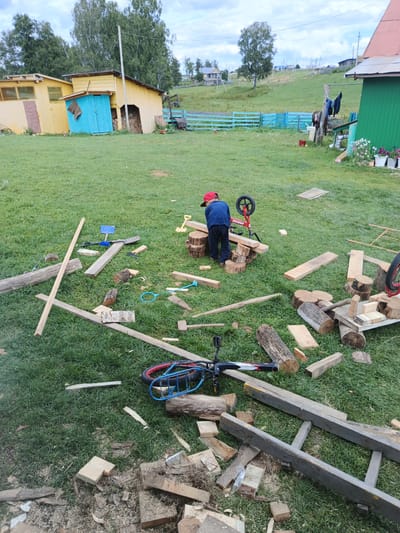 In the rural settlement of Islambaevo, Russia, a young boy wearing a red cap and a blue shirt is observed in the foreground of the image. He is standing amidst a collection of lumber and logs scattered across a grassy yard. The boy appears to be engaged in an activity involving these materials, with a small red bicycle positioned behind him, its front wheel slightly elevated. A wooden saw is also visible near the boy, suggesting he might be cutting or manipulating the wood.
The environment includes a wooden shed with a blue door and a yellow exterior in the background, along with other structures and fencing. Trees are present in the distance, contributing to a rural landscape. The ground is covered in green grass, with visible sawdust and wood chips scattered around the immediate area of the boy and the lumber, indicating recent woodworking activity. The overall scene depicts a rustic outdoor setting with evidence of construction or repair work in progress.