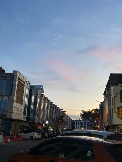 This media file captures a street scene in Pekanbaru, Indonesia, during what appears to be twilight. The perspective is a low-angle shot looking up and to the right, with the buildings and sky occupying most of the frame.
The subjects are primarily buildings and vehicles. A row of multi-story buildings with a mixture of architectural styles lines the right side of the image. Some of these buildings feature decorative facades and large windows, while others appear more modern. The buildings are lit from within, suggesting it is evening. Several cars are parked along the street, including a prominent orange vehicle in the foreground on the far right, and a white van further down the street. Some traffic cones are visible, possibly indicating construction or a temporary road closure. No people are clearly visible in the frame, though they may be inside the buildings or obscured.
The sky is a significant element, displaying a gradient of colors from a deep blue on the left to a lighter, pale yellow-orange on the right, indicative of sunset or sunrise. Wispy clouds, tinged with pink, are scattered across the blue portion of the sky. A street lamp is illuminated, casting a warm glow.
The overall impression is one of a quiet street at the end of the day. The lighting and the soft colors in the sky create a somewhat tranquil atmosphere. There is no visible text within the image.