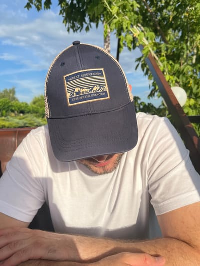 A man wearing a white t-shirt and a dark blue baseball cap is seated outdoors. The cap has a patch with a mountain landscape design and the text "GREAT MOUNTAINS" and "EXPLORE THE UNKNOWN." The man's face is mostly obscured by the cap. He has a beard. Sunlight casts shadows on his t-shirt, suggesting it is daytime. Green foliage and a blue sky with clouds are visible in the background, indicating an outdoor setting, possibly a park or a garden. The man's arm is resting on a surface, and his hand is visible. The scene appears relaxed and casual.