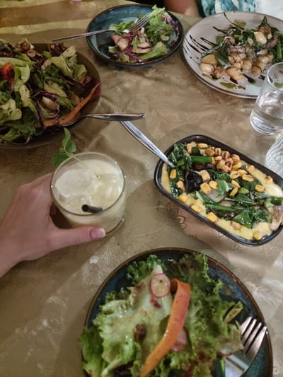 A table laden with various food dishes is seen from an overhead perspective. Dishes include salads, gnocchi with dark sauce drizzle, a corn and greens dish with a creamy yellow base, and another salad on a smaller plate. Glassware, including a water carafe and glasses, are present. A hand is holding a glass with a light-colored beverage, ice, and a straw. The table is covered with a patterned, light-colored cloth. The location is suggested as Mitzpe Ramon, Israel.