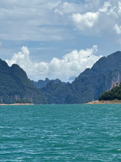 The image displays a serene natural landscape, dominated by towering, densely forested mountains that rise dramatically from a tranquil turquoise body of water. The mountains, covered in lush green foliage, have a rugged, uneven texture, with multiple peaks and ridges visible. The water's surface is slightly rippled, reflecting the sunlight and creating a shimmering effect. Along the shoreline where the mountains meet the water, there is a band of dry, sandy earth, hinting at a potentially lower water level or a distinct geological feature.

The sky above is a bright, clear blue, punctuated by fluffy white clouds that are scattered across the frame. The lighting suggests it is daytime, likely mid-morning or afternoon, with the sun casting a gentle illumination on the scene. The overall atmosphere is peaceful and majestic, evoking a sense of natural beauty and remoteness.

The scene is consistent with the geography of Ban Chiao Lan, Thailand, known for its dramatic karst mountains and the Cheow Lan Lake, which is part of the Khao Sok National Park. There is no activity or event taking place; the image captures a static, breathtaking view. No people or man-made objects are visible, emphasizing the untouched nature of the environment. The colors are vibrant, with the deep greens of the mountains contrasting with the bright blue of the sky and the rich blue-green of the water. There is no visible text in the image.