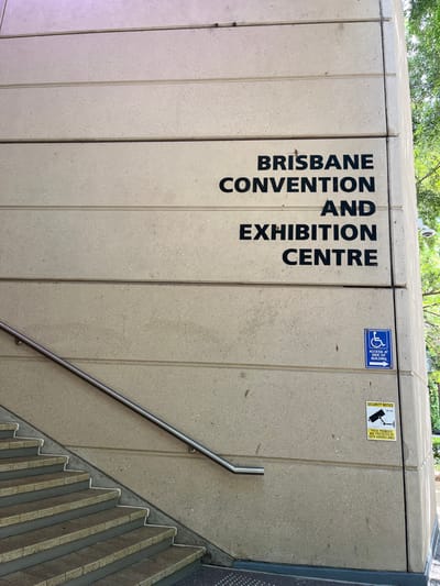 The image displays a light grey concrete wall, forming part of the exterior of a building. The wall features distinct vertical panel lines. Large, black, capitalized letters are painted vertically on the left side of the wall, spelling out "BRISBANE CONVENTION AND EXHIBITION CENTRE" from top to bottom.

To the right of this text, near the top edge of the wall, are two rectangular signs. The first, on the left, is blue with a white wheelchair accessibility symbol and the text "ACCESS AT -> ENTRY AT BUILDING". The second sign, to its right, is yellow with black text and a security camera icon, reading "SECURITY NOTICE THESE PREMISES ARE PROTECTED BY CCTV SURVEILLANCE". A physical security camera is also visible at the very top edge of the concrete structure, above these signs.

A polished metal handrail extends diagonally downwards and to the right, affixed to the wall. Further to the right, a staircase composed of concrete steps with textured treads descends. Green foliage from trees is visible along the top edge of the image, indicating an outdoor setting. The scene is brightly and evenly lit, suggesting daytime. No people or specific activities are depicted. The location is identifiable as the Brisbane Convention and Exhibition Centre in South Brisbane, Australia.