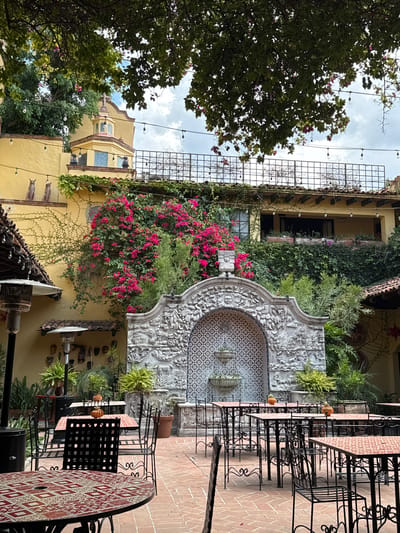 The image captures a scene in San Miguel de Allende, Mexico, in an outdoor courtyard setting. A stone fountain with intricate carvings and blue tile accents serves as a focal point. Red bougainvillea vines drape down the adjacent wall of a yellow building. Black wrought iron tables and chairs are positioned on a brick patio. Green trees and foliage are present, adding a natural element to the urban environment. Decorative items adorn the walls, contributing to the cultural and artistic ambiance. Overhead lights are strung across the courtyard.