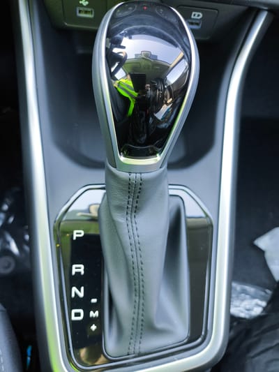 The image is a close-up, vertical, eye-level shot of an automatic car gear shifter. The shifter itself is a sleek, modern design with a leather boot and a highly polished, reflective top. The gear selection indicator is illuminated, showing the "P" (Park) position. The background is dark and out of focus, but appears to be the interior of a car, possibly with some light reflecting off surfaces. The location of Volzhskiy Utes, Russia is not visible or suggested by anything in the image.