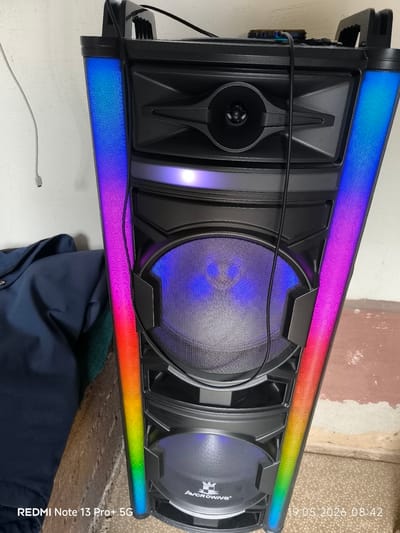 The image shows a black, multi-tiered speaker with rainbow-colored LED lights running vertically along its edges. The speaker has multiple circular grills over the internal components. "AVCRROWNS" is printed near the bottom. A black cord runs from the top of the speaker. The speaker is positioned near a white wall and a section of stained, light-colored flooring. Part of a blue garment is visible on the left. The image was taken on May 19, 2026, at 08:42. The phone that took the picture is a Redmi Note 13 Pro+ 5G. The location of the image appears to be in Pakhtakor, Uzbekistan.