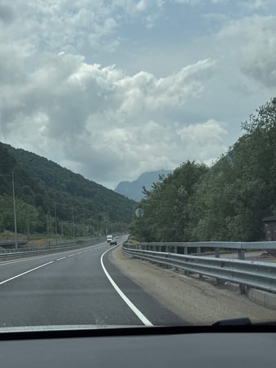 A highway road is pictured in the city of Chvizhepse, Russia. The road is made of asphalt and has white lines to mark the lanes. On the right side of the road is a metal guardrail. Beyond the guardrail is a forest with green trees. On the left side of the road, there is a steep, tree-covered mountain. The sky is cloudy and gray. There are a few cars on the road. The image appears to have been taken from inside a vehicle.