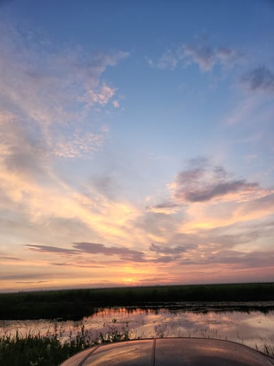 The image shows a vertical, panoramic view of a sunset over a body of water, likely in Atameken, Kazakhstan. The sky is filled with dramatic clouds painted in hues of orange, pink, and purple, transitioning into a clear blue at the top of the frame. The setting sun casts a warm glow, illuminating the undersides of the clouds and reflecting on the still water.

The water's surface acts as a mirror, doubling the vibrant colors of the sky and creating a serene, almost surreal landscape. Along the edge of the water, there is a strip of dark green vegetation, possibly reeds or tall grass, adding texture and contrast to the scene. To the right of the frame, the curved, metallic surface of an unidentified object is visible, reflecting the colors of the sunset.

The time of day is clearly sunset, given the intense colors in the sky and the soft light. The weather appears calm, with still water suggesting little to no wind. The location cues, such as the flat, open landscape and the presence of water and vegetation, are consistent with a rural or natural setting. The overall mood of the image is peaceful and awe-inspiring, evoking a sense of natural beauty and tranquility. There is no visible text in the image.