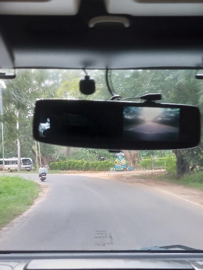 The image depicts a scene from inside a vehicle on a road in the city of Kisanga, Tanzania, viewed through the windshield. In the immediate foreground, a dark gray vehicle interior roof lining is visible, along with an overhead console and a centrally mounted rearview mirror. A dashcam or similar device is attached to the windshield below the roof lining, with its wires visible. The rearview mirror incorporates a small screen on its right side, displaying a forward-facing camera view of a sunlit, tree-lined road.
Beyond the windshield, a two-lane asphalt road curves gently to the left into the mid-ground. The road surface appears dry. On the right side of the road, a male individual is operating a motorcycle, traveling away from the camera. The motorcycle is heavily laden with a tall, bulky package secured to its rear, obscuring much of the rider. The road features dirt and grass shoulders on both sides.
Further down the road, on the left, a white minibus with a blue stripe is parked or stationary on the shoulder. Its front destination board is partially visible. On the right side of the road, past the curve, a prominent blue and yellow circular crest or emblem, consistent with a municipal or institutional sign, is displayed amidst green foliage. Tall trees line both sides of the road, creating a dense green environment. A utility pole is visible on the far left. The overall scene is consistent with a daylight setting in a semi-urban or peri-urban area.