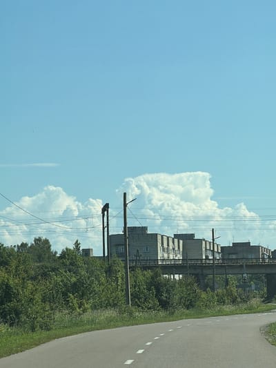 The image captures a scene from a road in Dobrivliany, Ukraine, likely on a sunny day with a clear blue sky and scattered cumulus clouds. In the foreground, a paved road curves to the right, marked with white dashed lines. Lush green vegetation, including trees and tall grass, lines the side of the road.

In the mid-ground, a concrete overpass spans across the scene, carrying what appears to be a railway line. Beyond the overpass, several multi-story apartment buildings with light-colored facades are visible, arranged in a row. Power lines are strung across the upper portion of the image, supported by utility poles. One pole in the middle ground has a dark, angular structure attached to its top. The overall impression is one of a residential area on the outskirts of a town, with infrastructure supporting transportation.