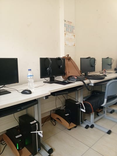 This image depicts a computer lab with multiple workstations, likely in a school or office setting. The room has beige walls and a tiled floor. Several black computer monitors are visible, some with headphones placed on top. Desks are made of light-colored laminate with metal supports. On one desk, a bottle of water and a computer mouse are present. Underneath the desks, several computer towers and power supply units are visible, some with illuminated green lights. A brown backpack leans against a monitor on one of the workstations. A grey office chair with a black bag on its seat is positioned in the foreground. The scene appears to be tidy and functional, suggesting a place of learning or work. The location is identified as Chutula, Mozambique.